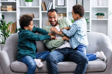 Evde babasıyla oynarken kanepede gıdıklanan neşeli küçük çocuklar. Mutlu siyah aile evde iyi hafta sonları.