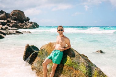 Young man in swim shorts stand near rocks beach of the tropical island Praslin Seychelles with a blue sky and white clouds.