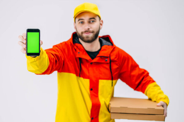 Smiling delivery worker with yellow thermal backpack showing smartphone, isolated on white background. Man using phone with blank green screen.