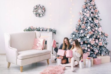 Gülümseyen genç kadınlar evdeki güzel Noel ağacının yanında bir hediye kutusu açıyor. Noel ve yeni yıl hediyeleri. Şömineli ve kanepeli beyaz oda..