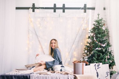 Yatakta, çelenklerin arkasındaki yastıkların arasında oturan genç kız. Yatağın yanında Noel ağacı.