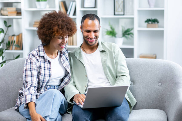 Couple Use Laptop Computer, while Sitting in the Living room of their Apartment. Boyfriend and Girlfriend Talk, Shop on Internet, Choose Product to Order Online, Watch Streaming Service