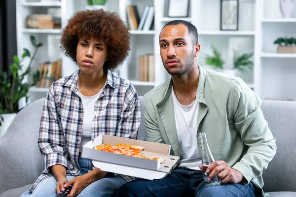Young multiethnic couple with serious facial expressions resting on sofa with pizza and drink and watching some thriller. Loving partners dressed in casual clothes spending weekend together indoors