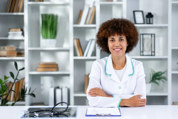 Portrait Of Smiling Female Doctor Wearing White Coat With In Hospital Office. African american physician or cardiologist in white lab coat uniform sitting at desk in her modern office