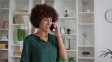 Mutlu Afro-Amerikan kıvırcık kadın portresi evde akıllı telefondan konuşuyor.