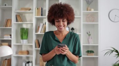 Akıllı telefon kullanan ve evde gülümseyen mutlu Afro-Amerikan kıvırcık kadın portresi. 