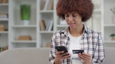 Ekose gömlekli Afro-Amerikan kıvırcık kadın portresi. Kredi kartıyla ödemeyi yapmak için akıllı telefon kullanıyor. Kanepede oturuyor.
