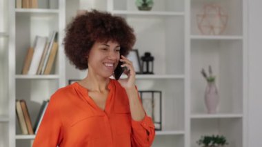 Afro-Amerikan kadın portresi kıvırcık saçlı, akıllı telefondan konuşan ve ofiste gülümseyen. 