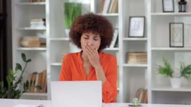 Ofiste dizüstü bilgisayarla çalışırken esneyen kıvırcık saçlı Afro-Amerikan kadın portresi. Uykusu var, yorgun, çok çalışıyor. Sıkıcı monoton iş konsepti. 