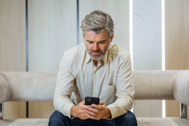 Lüks modern ofisteki koltukta oturan resmi giysili kıdemli bir adam telefon kullanıyor.