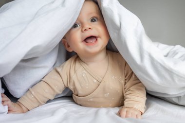 Gülen bebek beyaz çarşafın altında yatıyor.