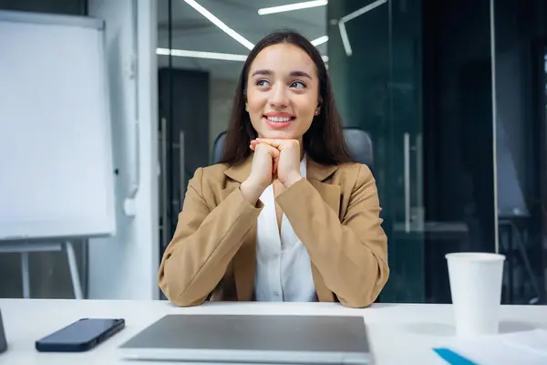 Young businesswoman reliving happy memories during a coffee break sitting at her desk in the office looking to the side with a smile of contentment and pleasure