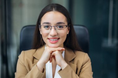 Gözlüklü, gülümseyen, kendine güvenen iş kadını kameraya bakıyor, çalışma masasında oturuyor. Mutlu düşünceli kadın serbest çalışan, iyi bir gelecek hayal eden, yeni iş fırsatları hayal eden. Elini çenenin altında tut.