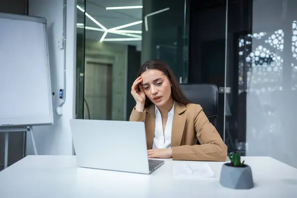 Tired female manager with dark hair sitting at office desk with laptop and grimacing from pain. Young lady wearing business suit having severe migraine during working process indoors.