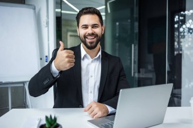 Resmi takım elbiseli yakışıklı sakallı bir adamın portresi modern ofiste gülümseyip el hareketi yapıyor.