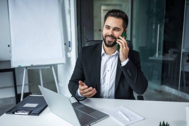 Gülümseyen işadamı telefonda konuşuyor, dizüstü bilgisayarla masada oturuyor, dost bir yönetici müşteriye telefonla danışıyor.