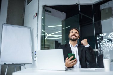 Neşeli genç sakallı adam modern ofiste masasında oturuyor hücre ekranında zafer mesajının tadını çıkarıyor, maaş mesajları alıyor..