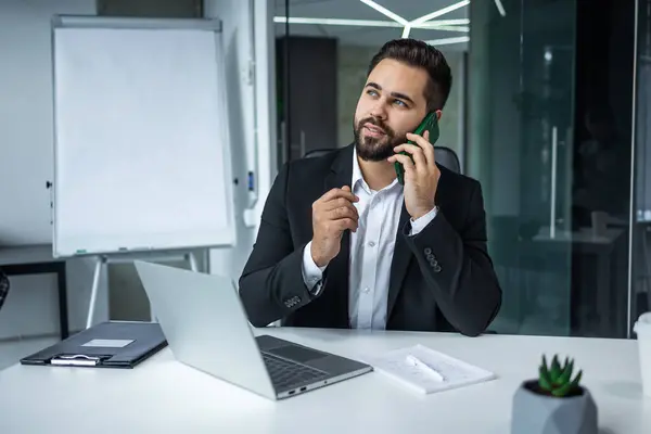 一个英俊的生意人在办公室里一边用电脑一边用手机聊天 免版税图库照片
