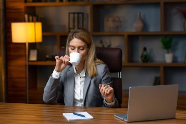 Nice coffee! Young beautiful woman holding coffee cup and keeping eyes closed while sitting at her working place