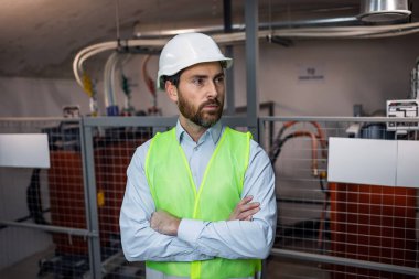 male engineer technician standing in technical room of electrical switchboard, wearing green safety vest, helmet in environment of electrical substation.