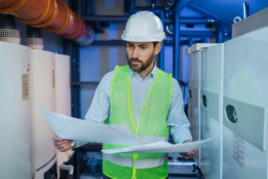 Focused engineer standing near heating system and checking correspondence of data with construction drawing.