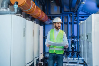 Bearded male engineer in helmet and vest inspecting manufactured equipment at factory heating room.