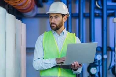 Heating plant technician standing by gas pipes and maintaining temperature inside power plant boiler room.