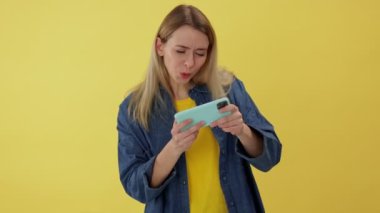 Duygusal ve komik kadın modern cep telefonuyla online video oyunları oynuyor. Sarı stüdyo arka planında izole edilmiş sürüş simülatörü olan akıllı telefon uygulaması kullanan neşeli, güzel bir kadın..