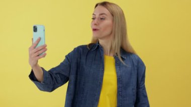 Sarışın, beyaz, hoş görünümlü bir kadın sarı stüdyo arka planından izole edilmiş modern cep telefonuyla görüntülü arama yapıyor.