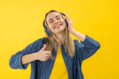 Sarı elbiseli, gülümseyen, 20 'li yaşlarda, müzik dinleyen kulaklıklı, sarı renkli duvar arkası stüdyo resminde baş parmağını gösteren genç bir kadın.