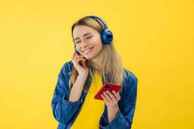 İnsanlar, müzik, duygu konsepti. Kaygısız kadın melodinin ritmiyle dans eder, gözlerini kapatır kulaklıkla yüksek sesle şarkı dinler, akıllı telefon tutar, eğlenir. Sarı renk arkaplanı