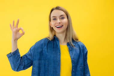 Sarı kazaklı, gülümseyen, eğlenceli genç bir kadın. Stüdyoda sarı duvar arkasında izole bir şekilde poz veriyor. Kameraya bak ve onaylayarak evet de. Tamam işaret işareti göstermen sorun değil.