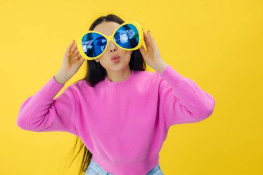 Büyük güneş gözlüklü çekici genç kadın sarı arka planda izole edilmiş şık pembe elbiseler giyiyor.