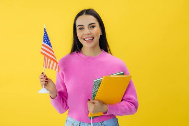 Elinde kitap tutan iyimser bir kadın ve sarı arka planla stüdyoda dururken ulusal bayramı destekleyen ve heyecanlanan Amerikan bayrağı..