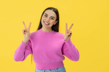 İki eliyle V işareti yapan genç bir kadın ve izole edilmiş sarı arka planda boş bir boşlukla gülümsüyor..