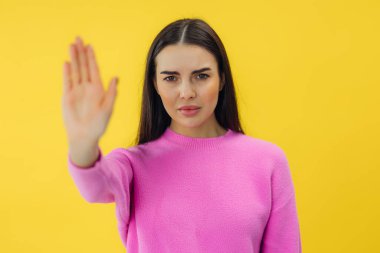 Ciddi genç bir kadın elini uzatmış, sarı arka planda izole edilmiş dur işareti yapıyor.