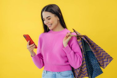 Çevrimiçi alışverişler için cep telefonunu kullanarak sarı arka planda izole edilmiş alışveriş torbaları taşıyan heyecanlı genç bir kadın.