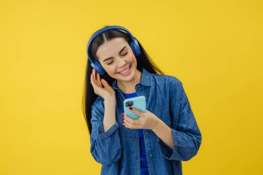 Elinde modern akıllı telefon ve kafasında kablosuz kulaklıkla neşeli bir şekilde dans eden ve en sevdiği şarkıyı dinleyen koyu renk saçlı genç bir kadın. Sarı stüdyo arka planında izole edilmiş..