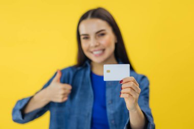 Neşeli esmer kadın baş parmağını gösteriyor ve elinde kredi kartıyla sarı arka plandan kameraya bakıyor. seçici odak