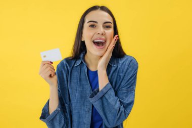 Heyecanlı çığlık atan genç bir kadın sarı arka planda tek başına duruyor. Elinde kredi kartıyla kameraya bakıyor..