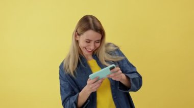 Duygusal olarak mutlu, gülümseyen bir kadın, sarı stüdyo arka planında izole edilmiş sürücü simülatörüyle modern akıllı telefon uygulamasında hevesle online video oyunları oynuyor..