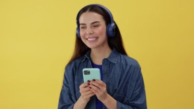 Kablosuz kulaklıklı neşeli beyaz kadın aktif müzik dinlerken ritm dansından ve telefondan keyif alıyor. Sarı stüdyo arka planında izole edilmiş..