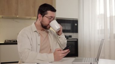 Mutlu gülümseyen adam akıllı telefon yazıcı mesajları kullanıyor, internet sosyal medya uygulamalarına göz atıyor, internette sohbet ediyor evde kahve içiyor ya da sıcak çay içiyor.