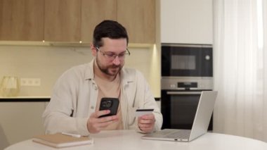 Mutlu adam internetten alışveriş yapıyor cep telefonunda kredi kartı numarasını yazıyor. Masasında oturuyor. Gülümseyen sakallı erkek satın alıyor, promosyonlar ve indirimlerle tatmin oluyor.
