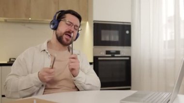 Neşeli adam modern bilgisayarla evde otururken kablosuz kulaklıkla müzikten zevk alıyor. Sanal davul çalmak için kalem kullanarak günlük giysiler giyen neşeli erkek meraklısı..