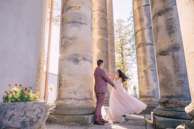 Zarif bir gelinlik içinde çekici bir gelin, sıcak bir yaz gününde, bir bahçede, taş sütunların yanında, bir damatla dans ediyor..