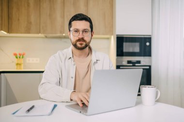 Laptop kullanan neşeli sakallı bir gencin portresi mutfak masasında oturmuş, uzaktan kumandayla uzaktan kumandaya bakıyor..