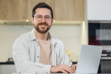 Gözlüklü neşeli sakallı adam evde uzaktan çalışırken modern laptop kullanıyor.. 