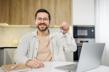 Yakışıklı, başarılı bir adam, kişisel işyerinde elinde gümüş anahtarlar olan, kablosuz dizüstü bilgisayarla oturuyor ve merkez ofisin arka planındaki kameraya bakıyor..