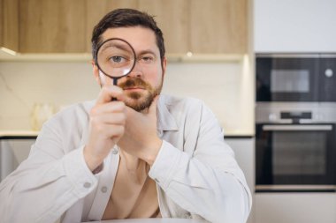 Büyüteçle bakan ve kameraya göz kırpan oynak yetişkin bir adamın portresi ve cilveli bir ifadeyle hareket eden kaşlar..